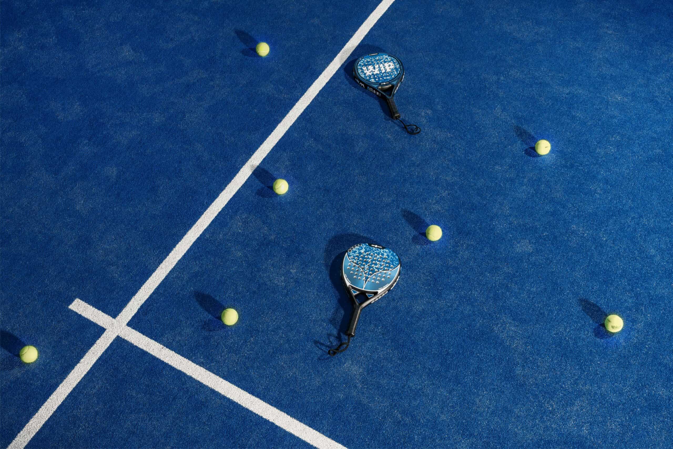 Zwei Padelschläger und mehrere gelbe Bälle liegen verstreut auf einem blauen Padelplatz. Weiße Linien unterteilen den Platz und bilden ein geometrisches Muster. Die Szene ist hell beleuchtet, was die leuchtenden Farben des Platzes und der Ausrüstung hervorhebt.