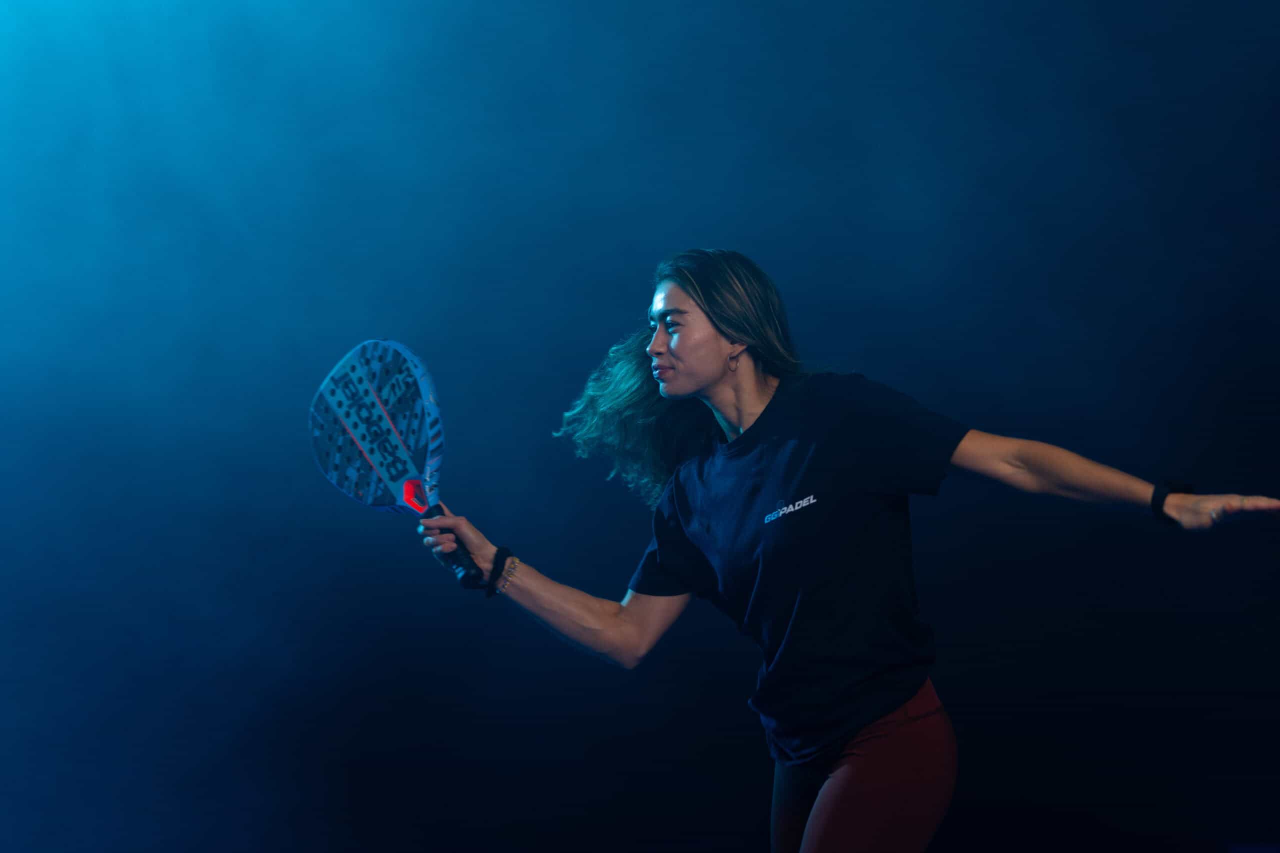Eine Frau spielt mit einem Paddle-Schläger und schwingt ihn durch die Luft. Ihr Haar weht hinter ihr her. Der Hintergrund ist dunkelblau und erzeugt eine dynamische und intensive Atmosphäre. Sie trägt ein schwarzes T-Shirt und dunkle Hosen.