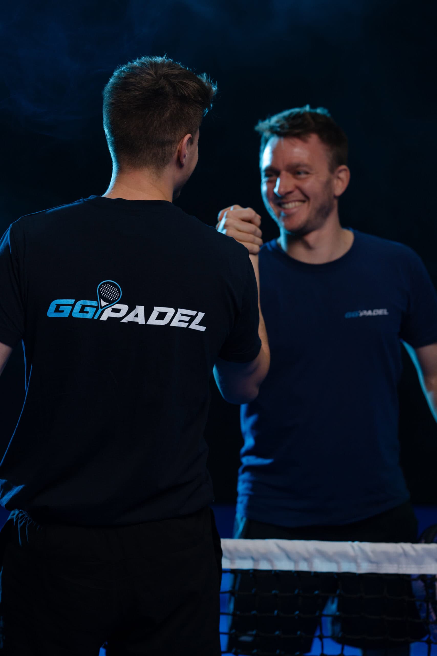 Zwei Männer stehen nachts auf einem Padelplatz und geben sich über dem Netz die Hand. Beide tragen dunkle Shirts mit GG Padel-Logos. Einer blickt in die Kamera, der andere mit dem Rücken. Sie lächeln und schaffen so eine freundliche Atmosphäre.