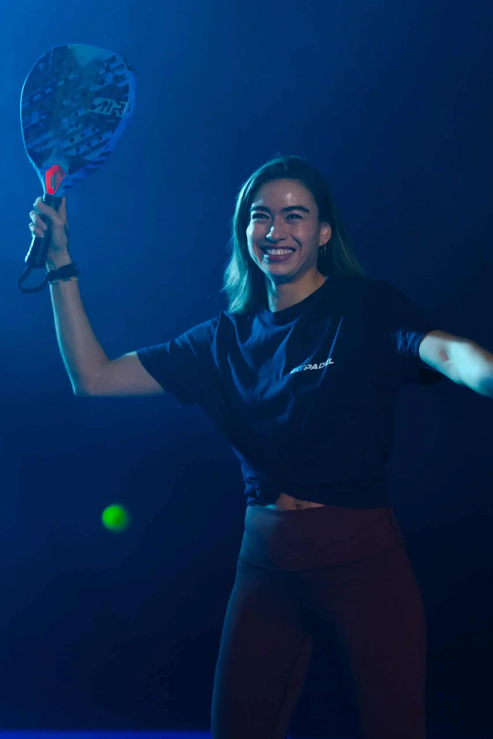 Eine Frau lächelt und spielt Paddle-Tennis in einer dunklen Umgebung. Sie hält einen Schläger in einer Hand und trägt legere Sportkleidung. Das Licht wirft einen blauen Ton über die Szene.