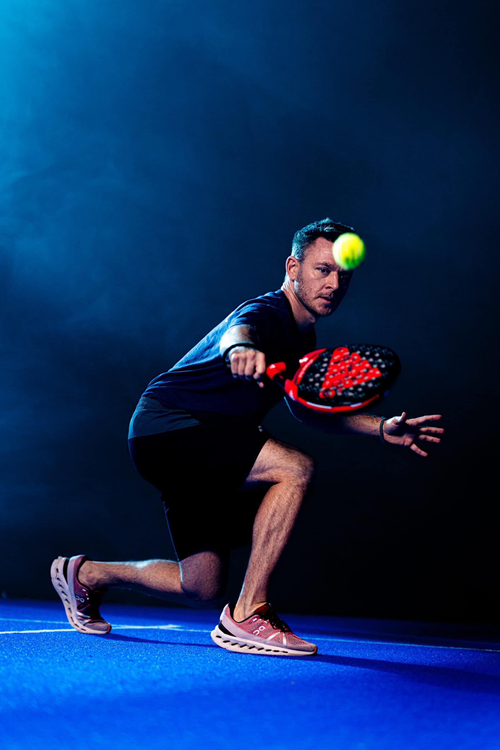 Eine Person in Sportkleidung stürzt sich auf einen blauen Padel-Platz und bereitet sich konzentriert darauf vor, einen gelben Ball mit einem roten Schläger zu schlagen. Der Hintergrund ist dunkel und ein stimmungsvoller Spotlight-Effekt hebt die Aktion hervor.