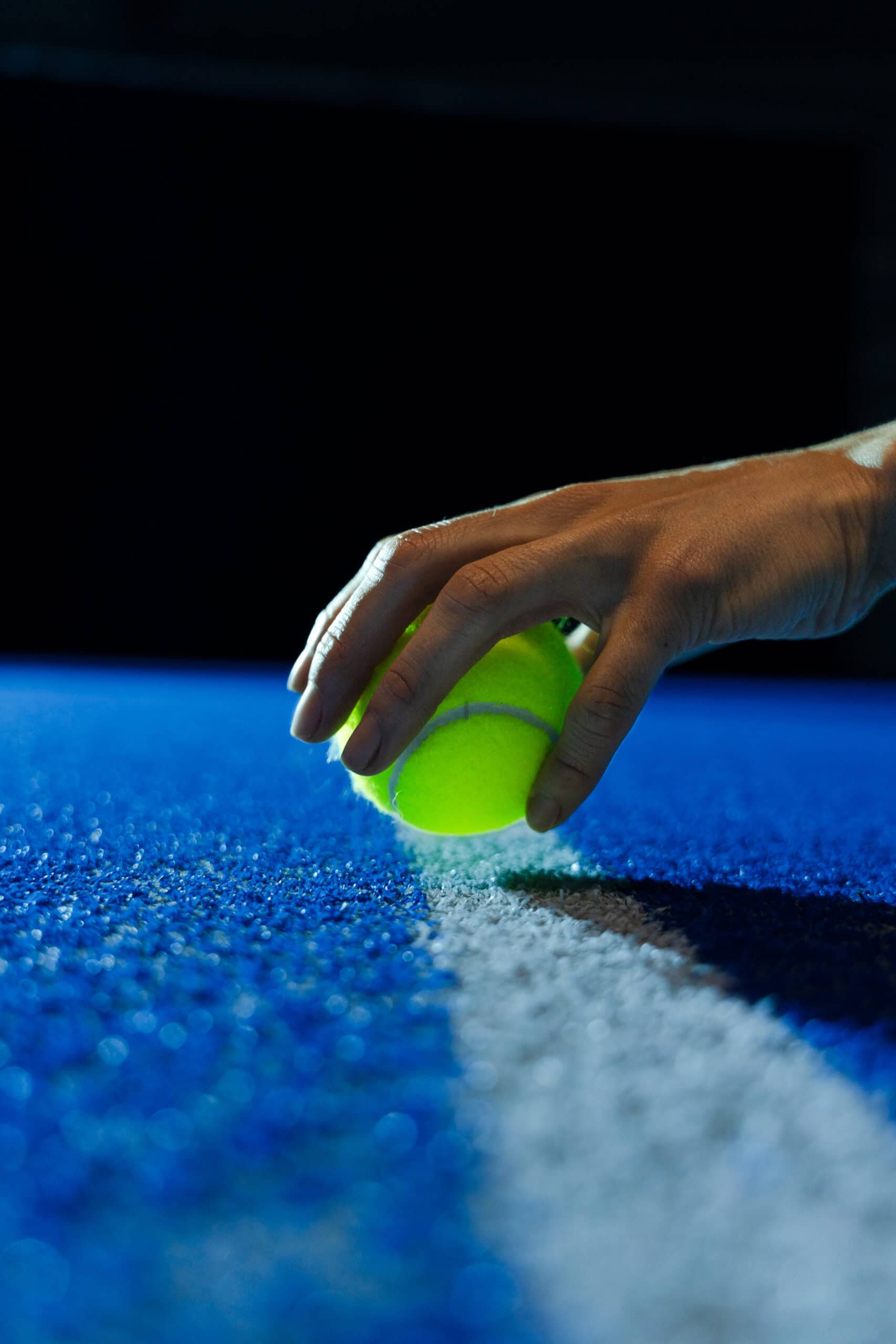 Eine Hand platziert einen gelben Tennisball unter dramatischer Beleuchtung auf einem strukturierten blauen Platz, nahe einer weißen Linie.